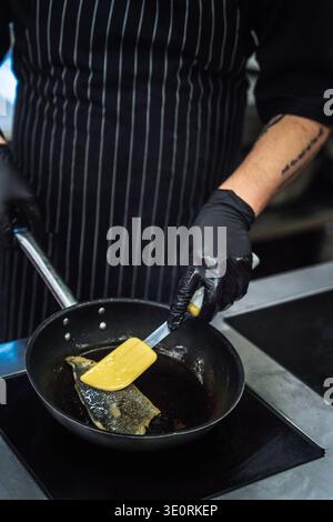 Eine Nahaufnahme der Hände eines Küchenchefs mit schwarzen Nitrilhandschuhen, mit einem gelben Spachtel ein Stück Fisch mit knuspriger Haut umwerfen. Der Koch trägt einen Stockfoto