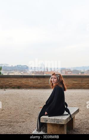 Junge rothaarige Frau auf einer Steinbank mit Panoramablick auf die Skyline von Rom und den Petersdom im Hintergrund. Ruhiger Reisemoment. Stockfoto