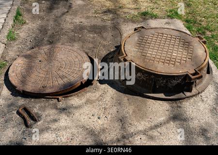 Rostiger offener Schächtenabdeckung und erhöhter Kanaldeckel auf gerissener Asphaltbegehfläche Stockfoto
