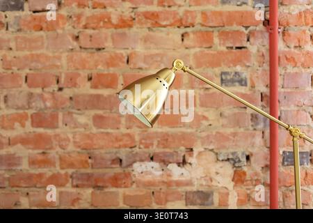 Moderne goldene Stehlampe vor einer roten Ziegelwand in einem Loftstil. Minimalistische Innenbeleuchtung mit industriellem Design und warmen Tönen Stockfoto