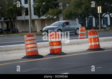 Straßensicherheitsfässer, die sich über eine frische Asphaltarbeitszone zwischen fahrenden Fahrspuren befinden, mit verschwommenen Fahrzeugen und Straßengebäuden dahinter Stockfoto