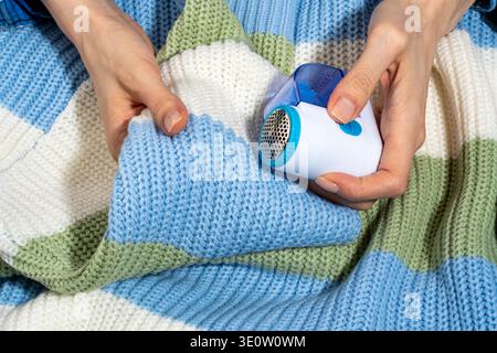 Weibliche Hände verwenden einen Stoffrasierer auf einem Strickpullover, um Fussel und Pilling vom Stoff zu entfernen. Konzept der Bekleidungsreparatur und Bekleidungspflege k Stockfoto