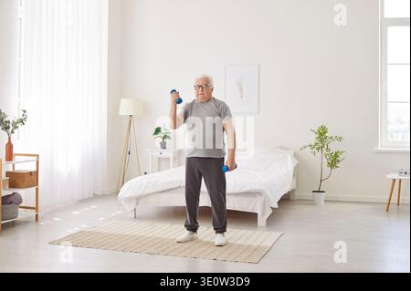 Fokussierter Seniorenmann hebt Kurzhanteln im Schlafzimmer, macht Morgenübungen, um aktiv und gesund zu bleiben. Stockfoto