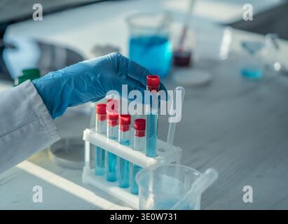 Nahaufnahme einer Hand mit Handschuhen, die ein mit blauer Lösung gefülltes Reagenzröhrchen zwischen mehreren Röhrchen in einem Rack hält. Laborglas und Pipette sind sichtbar und betonen wissenschaftliche Forschung und Innovation Stockfoto