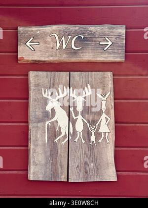 Rustikales hölzernes Schild mit WC (WC) mit Pfeilen nach links und rechts, montiert an einer roten Holzwand. Dekorative Tafeln zeigen ein illustriertes Tier Stockfoto