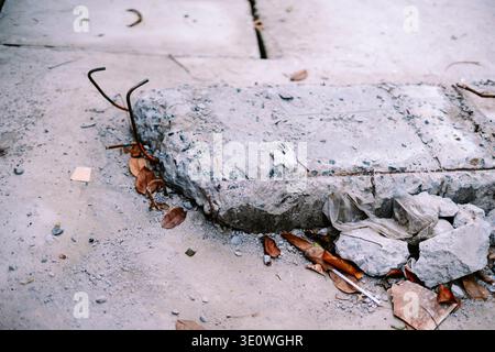 Nahaufnahme eines vernachlässigten Bereichs mit gebrochenen Betonplatten, freiliegendem rostfreiem Bewehrungsstahl, verstreuten trockenen Blättern und entsorgtem Kunststoff auf sandigem Boden, depic Stockfoto
