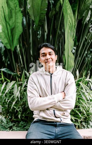 Ein lächelnder junger asiatischer Mann mit lässigem Kapuzenpullover posiert mit überkreuzten Armen vor dem Hintergrund lebendiger, grüner tropischer Pflanzen und strahlt eine freundliche und familiäre Atmosphäre aus Stockfoto