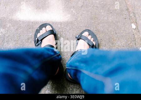 Die Füße einer Person in bequemen schwarzen Sandalen und blauen Jeans stehen auf einem strukturierten Betonpflaster, der aus der First-Person-Perspektive aufgenommen wurde Stockfoto