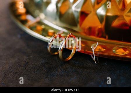 Zwei wunderschöne goldene Eheringe mit kleinen Diamanten ruhen auf einer dunklen, strukturierten Oberfläche. Ein glänzender, verzierter goldener Tablett sorgt für eine warme, reflektierende Ba Stockfoto