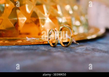 Elegante goldene Eheringe, die Liebe, Engagement und Zweisamkeit symbolisieren. In Nahaufnahme auf einer dunklen, strukturierten Oberfläche mit verschwommenem Gold Stockfoto