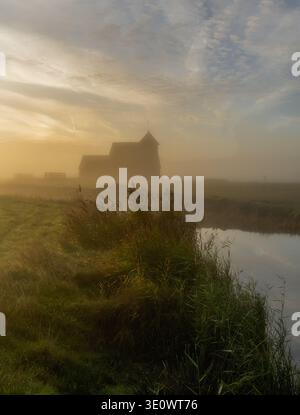 St. Thomas Becket (Fairfield) Kirche auf einem nebeligen Romney Marsh in Kent bei Sonnenaufgang mit Reflexionen im glatten Wasser des Puddledock Kanals Stockfoto