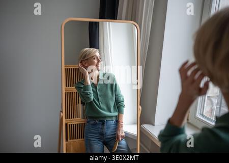 Lächelnde Frau in ausgefallener Kleidung, die sich im Spiegel spiegelt und mit positivem Ausdruck blickt Stockfoto