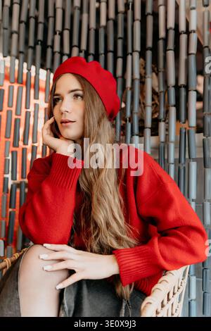 Frau Beret Fashion Junge Frau in einer roten Baskenmütze und Pullover sitzt nachdenklich vor einem dekorativen perlenbesetzten Vorhanghintergrund Stockfoto