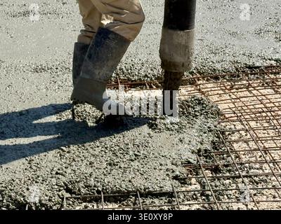 Nahaufnahme eines nassen Betons, der aus einem Pumpenschlauch auf ein Stahlbewehrungsgitter gegossen wird, mit den Stiefeln eines Bauarbeiters auf einer Baustelle. Stockfoto