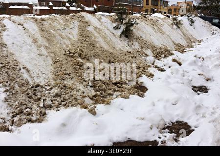 Wilde urbane Winterszene mit einer großen schmutzigen Schneebank am Straßenrand. Überreste der Schneeräumungsbemühungen der Stadt, die eine unordentliche Landschaft schaffen Stockfoto