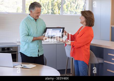 Reifer erwachsener Mann und ältere Frau mit gerahmtem Zertifikat, lächelnd in der Küche in der Nähe des Laptops Stockfoto