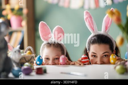 Fröhliches Mutter-Tochter-Duo in verspielten Hasenohren gucken über einen Tisch, der mit bunt bemalten Ostereiern bedeckt ist. Umgeben von Frühlingsblumen und festi Stockfoto