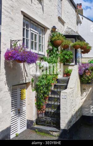 Charmantes weißes Cottage mit hängenden Blumenkörben, Topfpflanzen und schmiedeeisernem Tor auf Steinstufen in Cornwall, England. Stockfoto