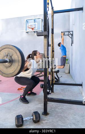 Eine Frau macht Squats mit einer Langhantel, während ein Mann in einem Fitnessstudio im Freien Pull-ups ausführt, um Kraft, Fitness und Hingabe zum Sport zu zeigen Stockfoto