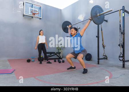 Ein konzentrierter männlicher Athlet führt Squats mit einer Langhantel aus, während eine weibliche Personal Trainer seine Technik in einem modernen Outdoor-Fitnessstudio beobachtet und fördert Stockfoto