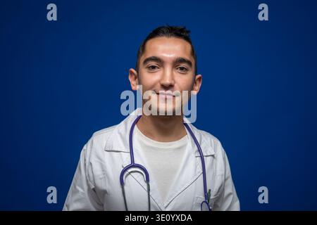 Junger Arzt mit weißem Mantel und lächelndem Stethoskop, stehend auf blauem Hintergrund, repräsentiert Gesundheit und Medizin Stockfoto