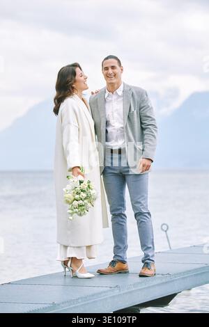 Glückliches Paar, das am Seeufer mit Berglandschaft steht, lächelnder Mann und Frau, die friedliche Momente in ruhigem Wasser genießen, während sie weiße Blüten halten Stockfoto