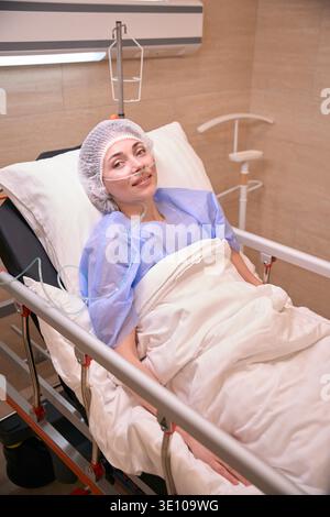 Lächelnde Frau in blauem medizinischem Kleid auf dem Krankenhausbett drinnen Stockfoto