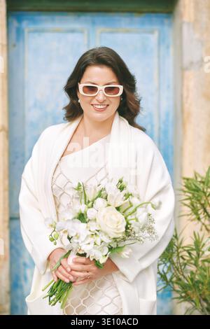 Lächelnde Braut mit weißem Hochzeitsstrauß, der neben der blauen Tür steht, elegante Frau in stilvollem Kleid und Sonnenbrille, die romantische Hochzeitstag-Porträt genießt Stockfoto
