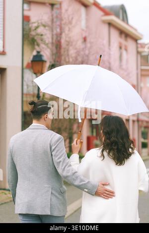 Romantisches Brautpaar, das zusammen unter weißem Regenschirm in leichtem Regen auf einer ruhigen Straße spaziert, Bräutigam hält Braut, während sie einen ruhigen, regnerischen Tagesspaziergang genießen Stockfoto