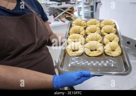 Baker trägt eine Schürze und blaue Schutzhandschuhe, hält ein Blechblech, gefüllt mit zwölf ungekochten Croissants, bereit zum Backen in einem Werbespot Stockfoto