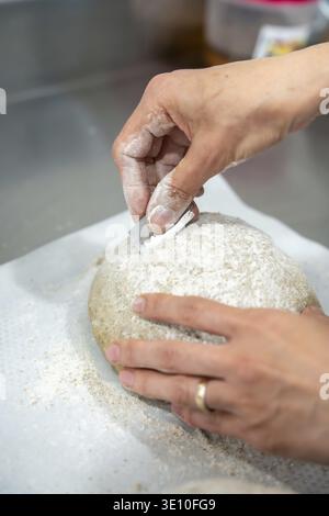 Backers Hände schneiden mit einer Rasierklinge einen runden Sauerteig, Formen die Kruste und bereiten den fermentierten Teig für das Backen in einer rustikalen Küche vor Stockfoto