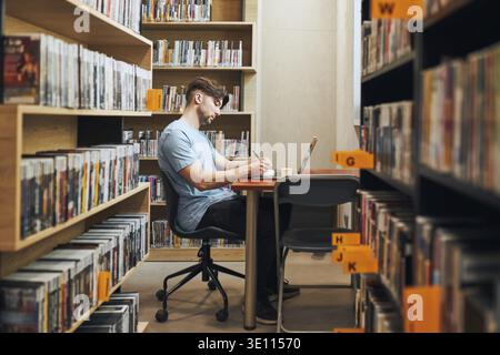 Schüler lernen in der Bibliothek. Junger Mann, der sich auf den Test am Laptop vorbereitet. Mann hört sich Online-Kurs an. Fokussierter Student, der für College-Prüfungen studiert. Verwenden Von Stockfoto