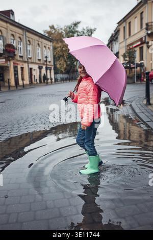 Kind mit einem großen rosa Regenschirm, der am regnerischen, düsteren Herbsttag durch die Pfütze in der Innenstadt läuft Stockfoto