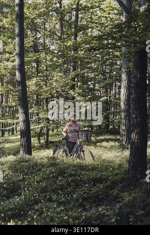 Aktive Frau, die freie Sommerferienzeit auf einer Fahrradtour in einem Wald verbringt. Frau trägt Fahrradhelm und Handschuhe halten Fahrrad mit Korb Stockfoto