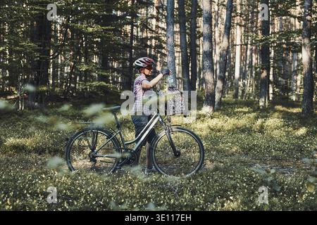 Aktive Frau verbringt freie Urlaubszeit auf einem Fahrradausflug in einem Wald an einem sonnigen Sommertag. Frau mit Fahrradhelm und Handschuhen, die das Fahrrad halten Stockfoto