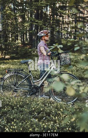 Aktive Frau verbringt freie Urlaubszeit auf einem Fahrradausflug in einem Wald an einem sonnigen Sommertag. Frau mit Fahrradhelm und Handschuhen, die das Fahrrad halten Stockfoto