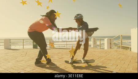 Ein gelehnter erwachsener Mann hält Kinderhände, ein Junge auf dem Skateboard am Meer, ein Korallenhemd-Helm Stockfoto