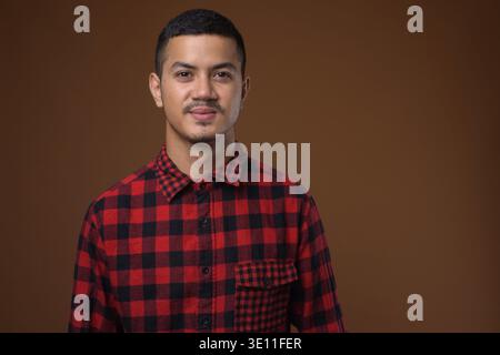 Studio shot Junger multi-ethnischen asiatischer Mann tragen rot kariertem Hemd gegen braunen Hintergrund Stockfoto