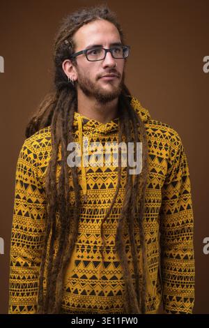 Studio shot von jungen hübschen Hispanic Mann mit Dreadlocks tragen Hoodie gegen braunen Hintergrund Stockfoto