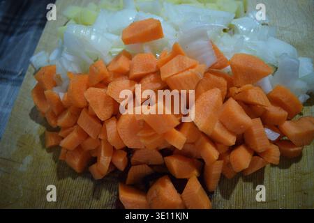 Eine detaillierte Hochwinkel-Nahaufnahme, die frisch gewürfelte rohe Kartoffeln, gehackte weiße Zwiebeln und leuchtend orangefarbene Karottenstücke zeigen, die auf einem Holzschneidebrett angeordnet sind Stockfoto