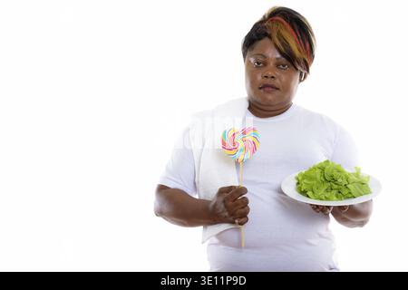 Studio Schuss von Fett schwarzen afrikanischen Frau hält Salat serviert Auf weißem Teller und herzförmigem Lollipop Stockfoto