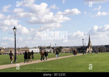 London, Großbritannien. März 2026. Wetter in Großbritannien. Menschen, die über die offene Weite von Blackheath spazieren, an einem hellen Frühlingstag ist der Turm der St. James's Church am Horizont sichtbar, flankiert von viktorianischen Laternenpfählen und historischen Gebäuden unter einem dramatischen blauen Himmel mit wehenden weißen Wolken. England. Credit:glosszoom/Alamy Live News Stockfoto