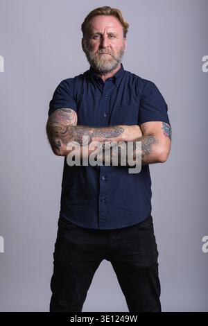 Studio shot reifer bärtiger Mann mit hand Tattoos stehend mit Waffen in grauer Hintergrund gekreuzt Stockfoto
