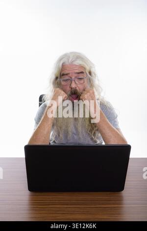 Studio shot der älteren bärtigen Mann Brille tragen, während Sie schockiert und mit Laptop auf hölzernen Tisch Stockfoto