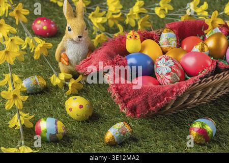 Ein Korb voller farbenfroher Ostereier und eine Kaninchenfigur mit gelben Blumen und Schokoladeneiern Stockfoto