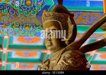 Nahaufnahme der traditionellen asiatischen Schutzstatue unter kunstvoller, farbenfroher Tempeldecke, ruhigem Gesicht und detailliertem Kopfschmuck, lebendig gemalten Mustern in Ba Stockfoto