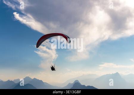 Die Silhouette des Solo-Gleitschirms steigt über den dramatischen blauen Himmel und die nebelige Bergkette darunter. Stockfoto