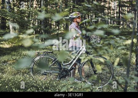 Aktive Frau verbringt freie Urlaubszeit auf einem Fahrradausflug in einem Wald an einem sonnigen Sommertag. Frau mit Fahrradhelm und Handschuhen, die das Fahrrad halten Stockfoto