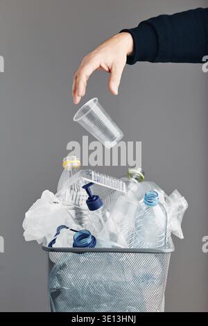 Männliche Hand wirft einen Plastikbecher in den Müll. Haufen von Plastikflaschen, Bechern und Beuteln, die zum Recycling in einem Metallbehälter gesammelt werden. Das Konzept der plastischen Verschmutzung A Stockfoto
