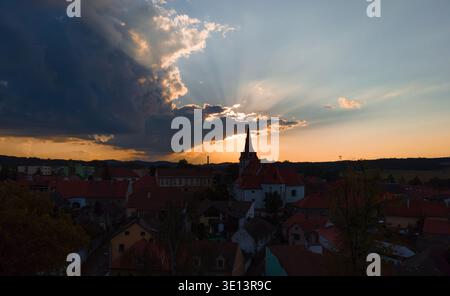 Die Kirche des Heiligen Wenzels in Netolice, die sich vor dem dramatischen Sonnenuntergangshimmel mit Sonnenstrahlen und Wolken über den historischen Dächern in Südböhmen, Tschechische Republik bildet Stockfoto